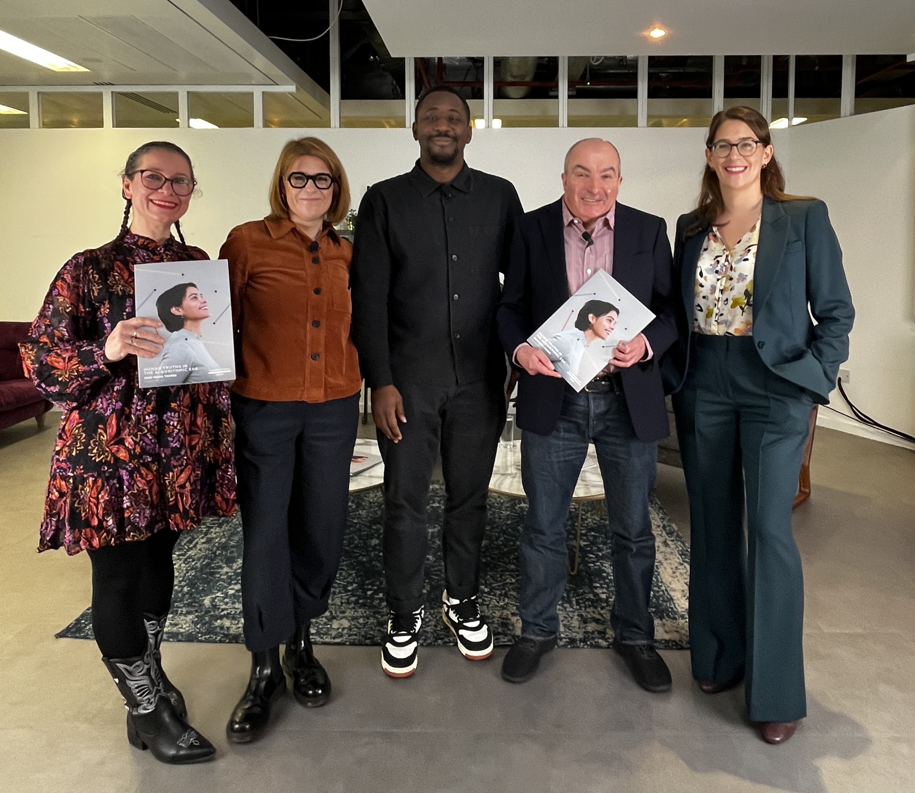 A group of five people standing together indoors, smiling at the camera. Two individuals on the left and one on the right are holding printed copies of a report featuring a person wearing headphones on the cover. They are dressed in smart and semi-formal clothing, standing in a modern studio-style setting with a rug, chairs, and soft lighting in the background.
